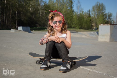 children's portraits, smiling little skater girl wearing sunglasses with retro hair and fake tattoo sleeve sitting on a skateboard in a skate park on a sunny day, children's photography, skater girl