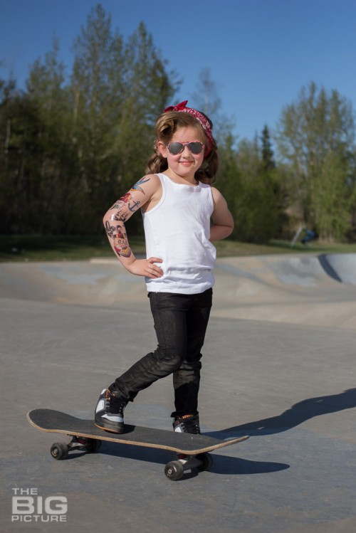 children's portraits, little skater girl smiling with sunglasses and retro hair and fake tattoo sleeve standing on a skateboard in a skate park on a sunny day, children's photography, skater girl