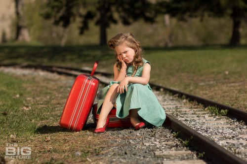 children's photography, young girl in red high heels sitting on a red suitcase by railroad tracks in a green dress and vintage hair, runaway